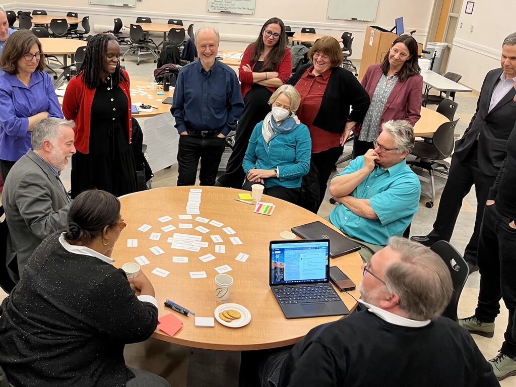A group of people in facemasks are standing around a round table with white HuMetricsHSS values cards organized in the shape of a tree with branches and roots made of values cards. This is a paper coffee cup placed on a round paper plate at the top of the tree with another round paper plate on top of the coffee cup with a set of values cards on the plate elevated above the table. A man on the right side of the table is pointing at the roots presenting the framework.