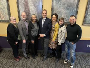 The HuMetricsHSS Team at the University of Washington standing together in front of a yellow wall with gray frames of artwork. From left to right is: Bonnie Russell, Jason Rhody, Bonnie Thornton Dill, Chris Long, Nicky Agate, Penny Weber, and Simo Sacchi.