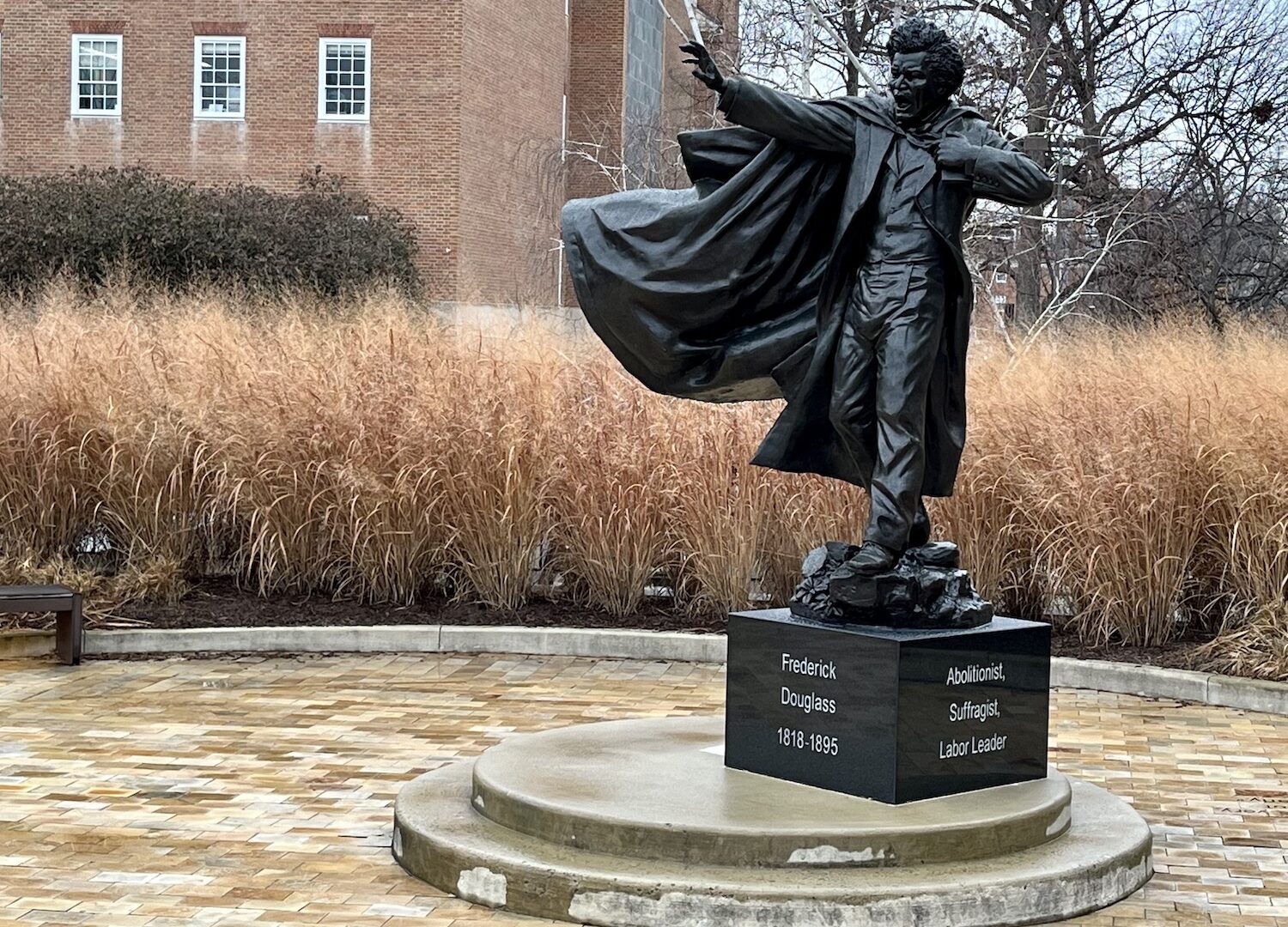 The Frederick Douglas statue on the University of Maryland campus depicts, as wikipedia puts it, "Douglass in the middle of a speech, with one arm outstretched, and a copy of his autobiography under the other arm." His mouth is open in a fiery speech. The statue is cast bronze on a black marble block with white text chiseled on front reading Frederick Douglas 1818-1895, on the side, white text is legible that reads: Abolitionist, Suffragist, Labor Leader. The figure is set off to the right of the image with a band of long, wheat colored grasses in the middle background, with three windows of the red brick colonial style building in the distant background.