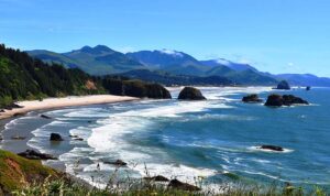 The Oregon coast. A sandy beach curving on the left with the Pacific Ocean gently crashing against it. There are dark rocks just off shore to the upper right, with mountains the background. The beach is surrounded by pine trees, standing majestically against the bright blue sky.